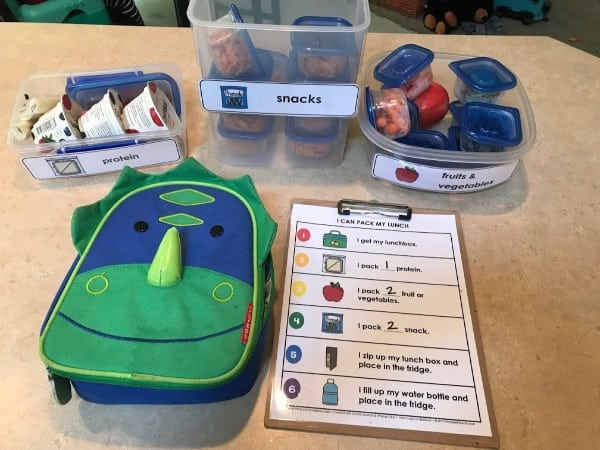 packing a school lunch with protein, snack, and fruit and vegetables on counter with a dinosaur lunchbox and printable chart