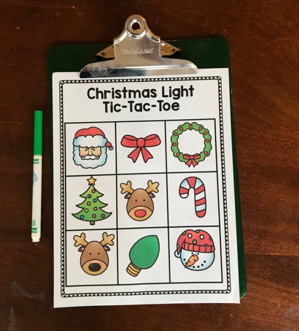 An easy christmas lights scavenger hunt printable on a green clipboard with a green marker. The printable reads Christmas Lights Tic-Tac-Toe and has cartoon pictures of Santa, a bow, wreath, tree, reindeer, candy cane, Rudolph, green light, and snowman.