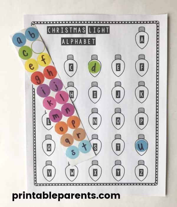 Christmas alphabet activity with 26 christmas lights with the capital letters of the alphabet on a black and white printable. Lowercase letters are written on color dot label stickers. The letters D and U on the printable Christmas alphabet worksheet are covered in dot stickers with lowercase letters.