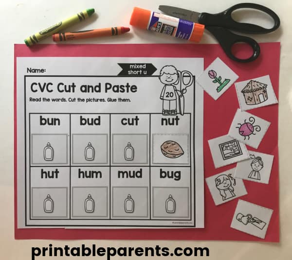 a white CVC cut and paste worksheet on a red construction paper background. There are seven little squares already cut out and scissors, a glue sticker, and a green and orange crayon.
