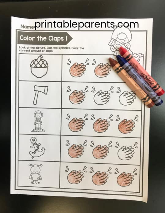 a phonemic awareness worksheet for syllables. It says Color the Claps 1 and has cartoon pictures of multiple syllable words. There is a brown, blue, and red crayon.